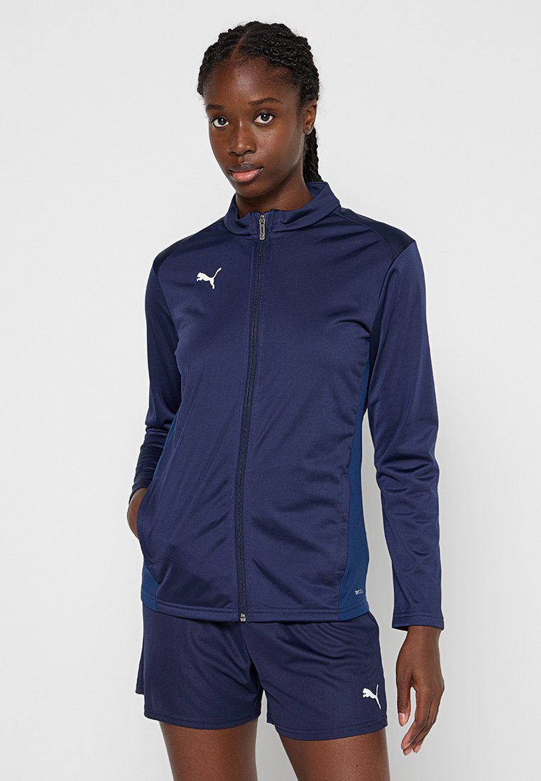 Chaqueta atlética navy con cremallera y cuello alto, mangas largas y logo de Puma, combinada con pantalones cortos a juego en un tono similar.