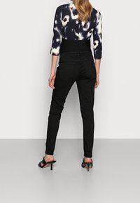 Mujer vestida con pantalones ajustados de tiro alto negros, sandalias de tacón negras y una blusa de patrón blanco y negro, de pie frente a un fondo liso.