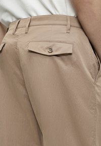 Gros plan d'une poche de pantalon beige avec rabat boutonné portée par une personne en chemise blanche, montrant les passants de ceinture et les détails des coutures latérales.