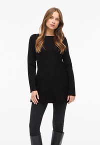 Zwarte tuniektrui met lange mouwen, rechte snit en ronde hals, gecombineerd met nauwsluitende zwarte broek en zwarte laarzen tot aan de knie.