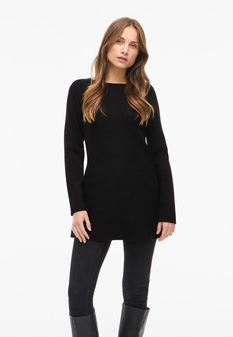Zwarte tuniektrui met lange mouwen, rechte snit en ronde hals, gecombineerd met nauwsluitende zwarte broek en zwarte laarzen tot aan de knie.