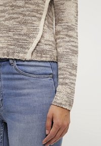 Maglione a righe beige e crema con orlo arrotondato e maniche lunghe, abbinato a jeans in denim blu con tasca frontale.