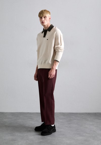 Jonge man met blond haar draagt een beige trui met een donkere kraag, burgundy broek en zwarte schoenen, staande tegen een effen grijze muur.