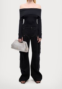 Person i svart off-shoulder långärmad topp och vida cargobyxor som håller en silverfärgad texturerad handväska, iklädd svarta öppna klackskor.