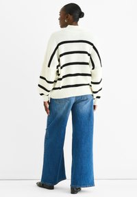 Maglione crema oversize con righe nere, texture a coste e spalle scese, abbinato a jeans denim blu a gamba larga e scarpe nere.