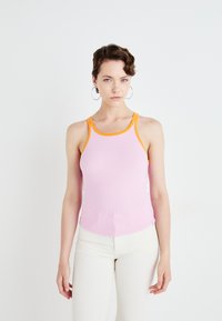 Geribbelde tanktop in zachtroze met contrasterende oranje bies. Getailleerd ontwerp met een licht gebogen zoom. Model draagt witte broek.