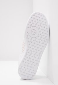 Biała podeszwa trampka z siatką bieżnika i okrągłym wzorem, z wytłoczonym logo "LACOSTE" w pobliżu pięty. But leży oparty o białą ścianę.