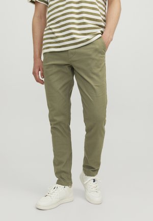 Pantaloni in cotone verde oliva con una vestibilità slim, dotati di tasche anteriori e una texture liscia. Indossati con una camicia a righe e sneakers bianche.