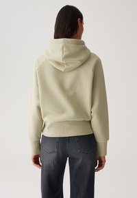 Persona che indossa una felpa con cappuccio beige chiaro e jeans grigi scuri, di spalle rispetto a uno sfondo chiaro e semplice.