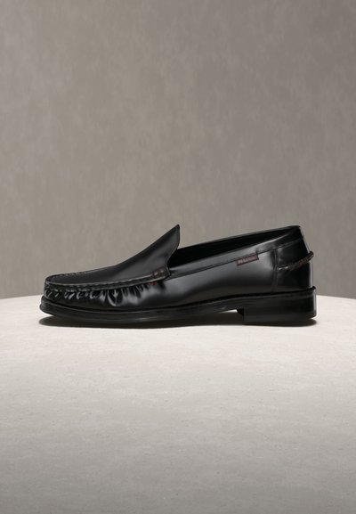 Mocassin noir en cuir à enfiler avec détails cousus et petit talon sur une surface et un fond neutres.