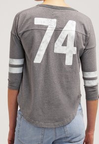 Grå långärmad tröja med vit "74" grafik och två vita ränder på varje ärm. Mjuk tyg, rundad nederkant, avslappnad design.