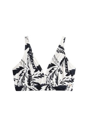 Mujer joven con bikini de estampado tropical en blanco y negro, de pie en una playa de arena con olas del océano y rocas al fondo.