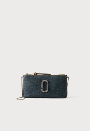 Petit porte-poignet rectangulaire en denim foncé avec fermeture éclair argentée, logo métallique à l'avant et une bandoulière chaîne argentée sur fond blanc.