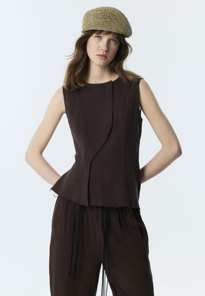 Jeune femme portant un haut sans manches marron foncé texturé et un pantalon avec un chapeau tissé beige, debout les mains derrière le dos sur fond blanc.