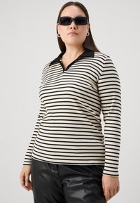 Camisa polo de manga larga a rayas en crema y negro con un cuello negro. El modelo lleva gafas de sol negras y está estilizado con pantalones negros brillantes.
