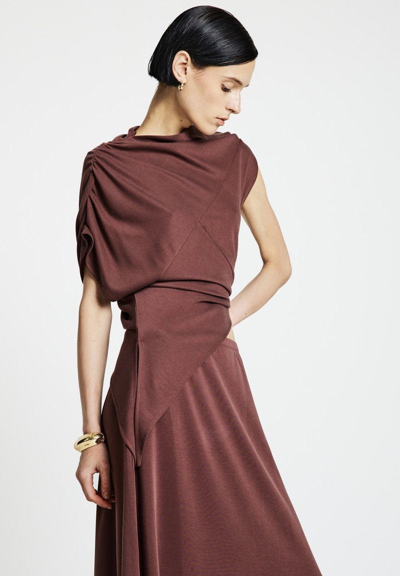 Robe marron drapée avec une coupe asymétrique et un tissu texturé. Design sans manches avec un détail froncé à l'épaule. Accent simple avec un bracelet.