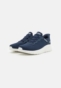 Scarpe sportive in rete di colore navy con suole in gomma bianca, con design slip-on e lacci elastici. Parte superiore testurizzata con dettaglio del logo del marchio.