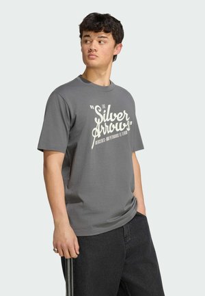 Młody mężczyzna w szarej koszulce z napisem „The Silver Arrows”, patrzący w bok, stojący na jednolitym jasnym tle.