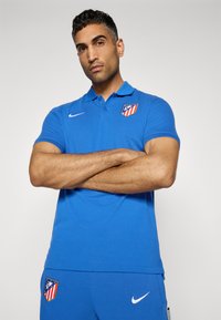 Blå polo-shirt lavet af et glat materiale, med krave, korte ærmer og et holdlogo på venstre bryst. Hvid Nike swoosh detalje.