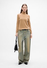 Beżowy sweter z golfem zestawiony z oversize'owymi niebieskimi dżinsami. Dżinsy mają wyblakły odcień. Modelka trzyma czarną torebkę. Czarnie buty dopełniają ten look.