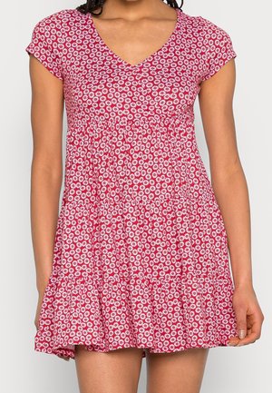 Femme portant une robe rouge à manches courtes avec un imprimé floral de marguerites blanches et un décolleté en V, vue des épaules jusqu'à la mi-cuisse.