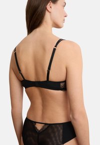 Frau von hinten gezeigt, die schwarze Unterwäsche mit Spitzenbesatz am BH-Bund und durchsichtigen Mesh-Pantys vor einem einfarbigen Hintergrund trägt.