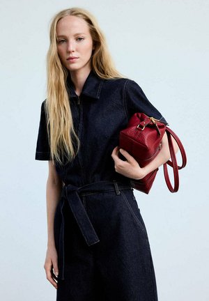 Femme aux longs cheveux blonds portant une combinaison en jean foncé, tenant un sac à main en cuir rouge sur un fond clair uni.