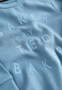 Svetlomodrá mikina s vystúpenou výšivkou názvu značky "TED BAKER" a slovom "BAKER" jemným písmom na prednej strane.