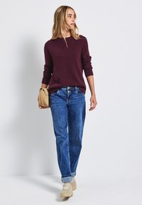Pull en maille bordeaux avec des poignets côtelés, associé à un jean droit bleu et des baskets beiges, portant un sac à main beige.