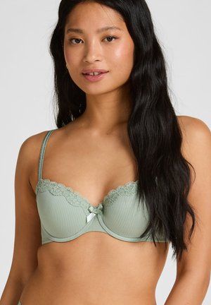 Frau mit langen schwarzen Haaren, die einen hellgrünen gerippten BH mit Spitzenbesatz und einer kleinen Schleife in der Mitte trägt und sanft lächelt.