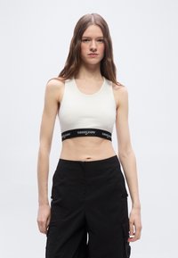 Νεαρή γυναίκα με μακριά καστανά μαλλιά, που φοράει λευκό crop top με μαύρη ταινία "tommy jeans" και μαύρο παντελόνι ψηλόμεσο, πάνω σε λευκό φόντο.