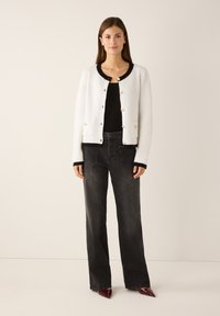 Witte gebreide cardigan met zwarte bies, voorzien van gouden knopen en zakken, gecombineerd met zwart hoogwaterjeans en puntige schoenen.