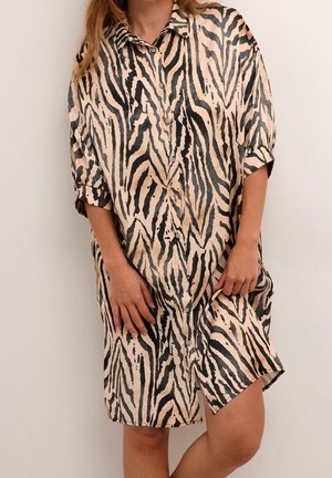 Frau trägt ein beige-schwarz gestreiftes knöpfenes Kleid mit Zebramuster und hochgekrempelten Ärmeln, steht vor einem schlichten hellen Hintergrund.