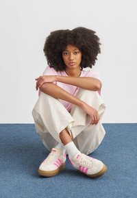 Beige joggesko med rosa striper, teksturert overdel og beige gummisåle. Lyserosa t-skjorte og beige vide bukser, sittende på et blått teppe.