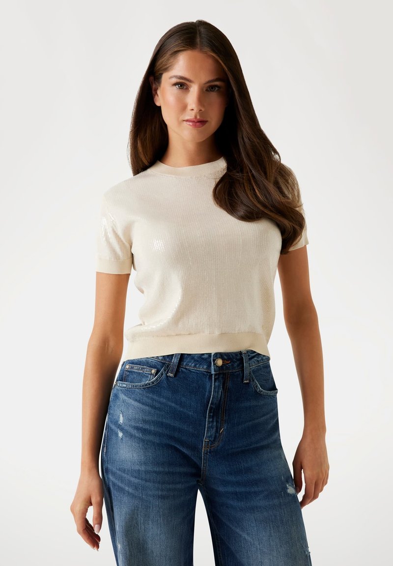Crème gebreide trui met korte mouwen en een subtiele glans, gecombineerd met mid-rise blauwe denim jeans met een lichte vervaging en verwerkte details.