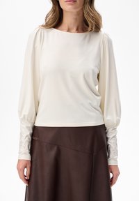 Frau, die eine cremefarbene Bluse mit langen Ärmel und gerafften Manschetten sowie Stickerei trägt, kombiniert mit einem dunkelbraunen Lederrock vor einem weißen Hintergrund.