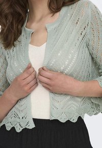 Světle zelený pletený cardigan s otevřeným vzorem, vlnitým lemem a rukávy ke třem čtvrtinám, vrstvený přes smetanový jemně strukturovaný top.