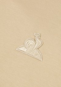 Tejido beige con un diseño de caracol bordado en blanco, que presenta una concha en espiral y acentos triangulares, centrado en el material.