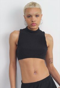 Zwarte geribbelde crop top met een hoge hals, gecombineerd met zwarte broek. Kenmerken zijn onder andere een navelpiercing en zichtbare tatoeages op de armen.