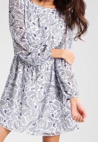 Mujer vestida con un vestido blanco de mangas largas y un patrón paisley azul, sosteniéndose un brazo con la otra mano.