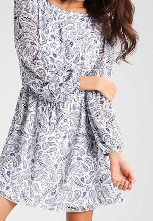 Kvinden bærer en hvid kjole med lange ærmer og et blåt paisley-mønster, mens hun holder den ene arm med den anden hånd.