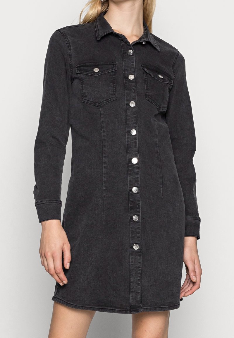 Femme portant une robe en jean noire boutonnée, manches longues, avec deux poches poitrine à boutons argentés.