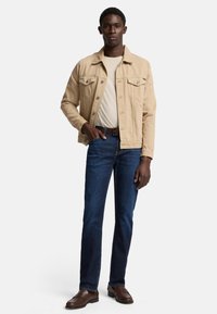 Beige Jeansjacke mit zwei Brusttaschen über einem hellbeigen Hemd, kombiniert mit blauen Jeans und braunen Lederloafers. Schlichte, taillierte Passform.