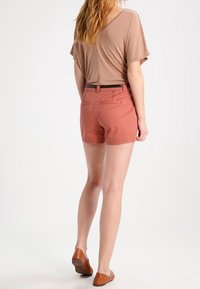 Brown loose-fitting top paired with peach shorts. The shorts feature a belted waist and back pockets. Worn with brown flat shoes.