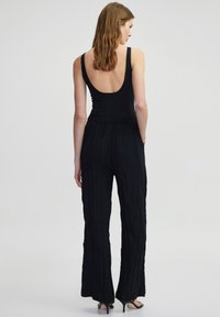 Zwarte mouwloze bodysuit met een lage rug, gecombineerd met zwarte wijde broek met textuur en verticale plooien. Hakken zijn zichtbaar.