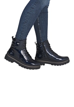 Schwarze, glänzende Knöchelstiefel mit Schnürung an der Vorderseite, seitlichem Reißverschluss und dicker Gummisohle, kombiniert mit dunkelblauen Jeans.