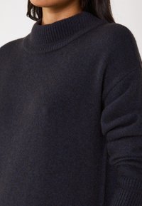 Maglione a maglia blu navy con colletto alto a coste, maniche lunghe e una texture morbida. Il tessuto presenta una profondità varia e sottile.