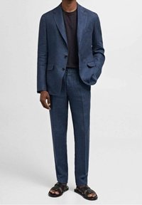 Completo navy su misura con finitura testurizzata, blazer aderente e pantaloni dritti, abbinato a una camicia scura e sandali neri.