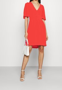 Rode jurk met een V-hals, korte pofmouwen en knoopdetails. Gecombineerd met een witte handtas en witte sandalen met bandjes. Soepele textuur.