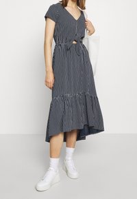 Vestito a righe navy con scollatura a V, maniche corte, dettaglio con fiocco davanti e orlo arricciato. Indossato con sneaker bianche e una borsa tote bianca.